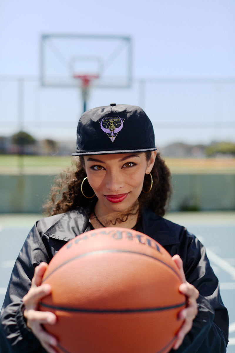 Woman in black Valhalla Hat with adjustable back and jacket holding a basketball on an outdoor court, basketball hoop visible in the background.