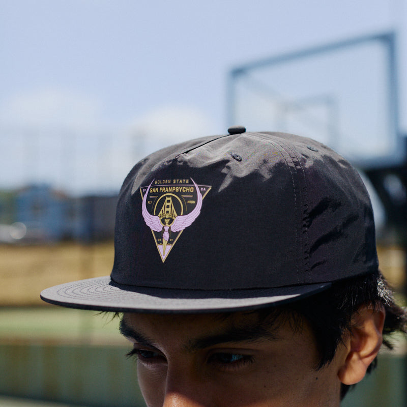 A young man wears a black Valhalla Hat with an adjustable back outdoors near a tennis or basketball court.