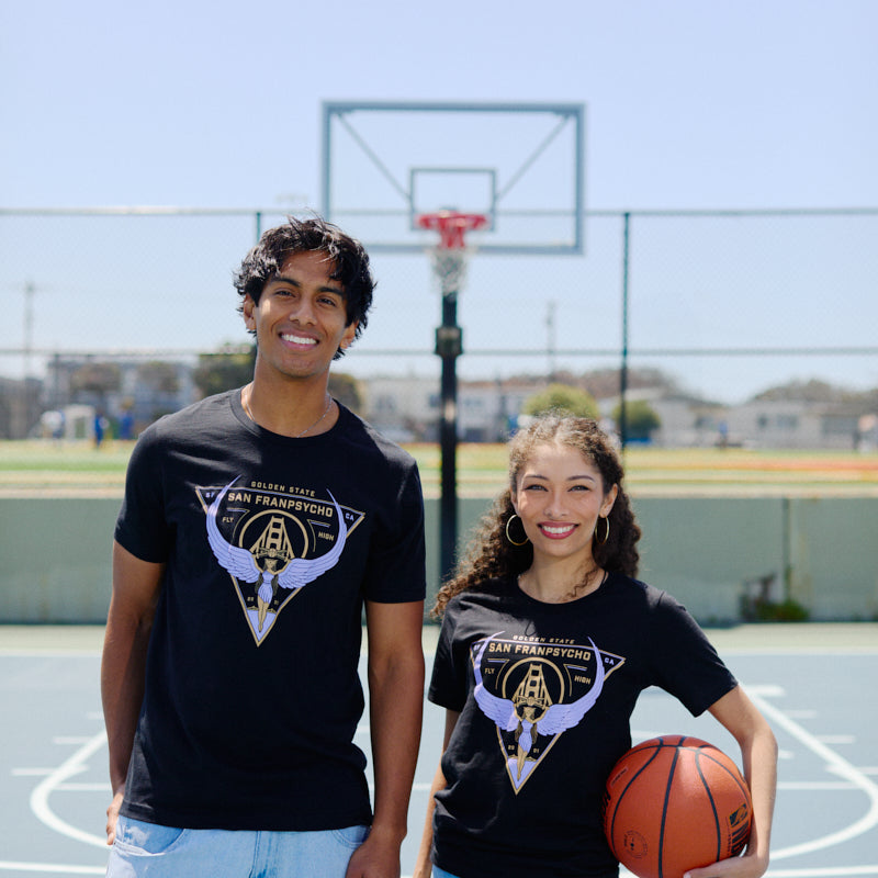 Two people smile on a basketball court in matching black Valhalla Tees, crafted from soft Airlume cotton; one holds a basketball, excited for the Valkyries season.