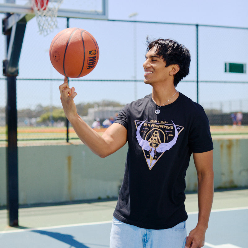 Smiling on an outdoor basketball court, a man wearing a Valhalla Tee spins a basketball on his finger.