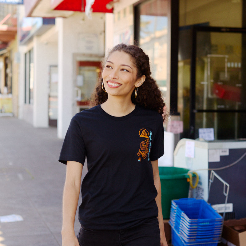 A woman wearing the Year of the Horse Tee walks on a sunny sidewalk lined with shops displaying Chinese signs, celebrating Lunar New Year.