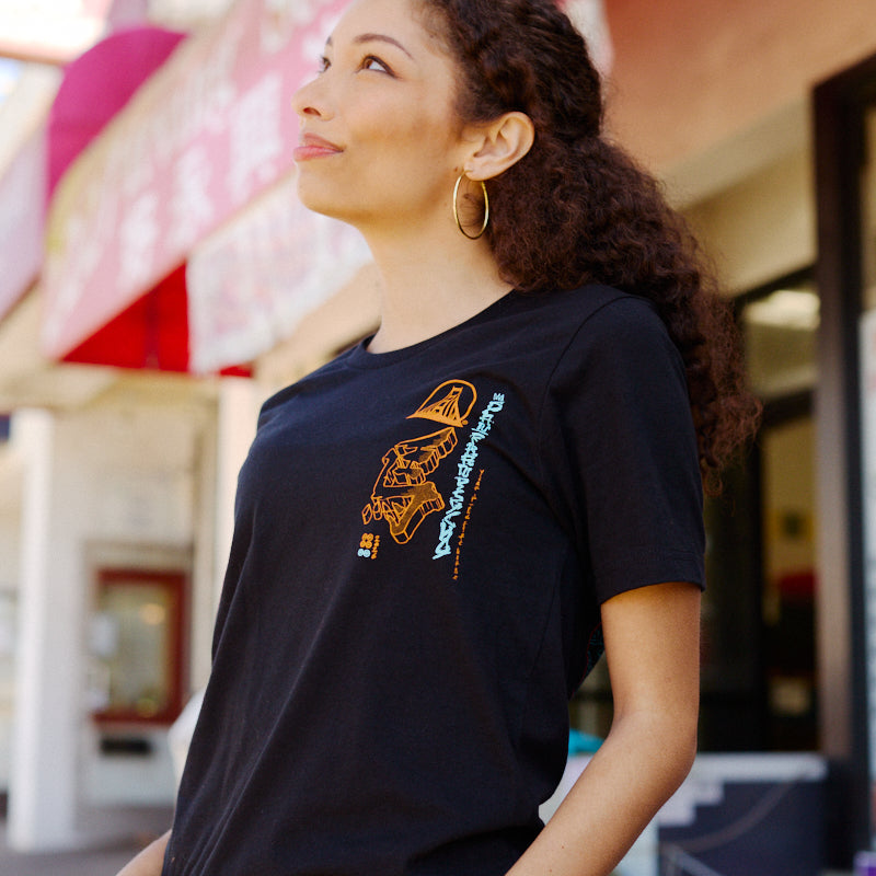 Wearing a Year of the Horse Tee, a woman stands outside among shops and festive signs, embracing the spirit of Lunar New Year.