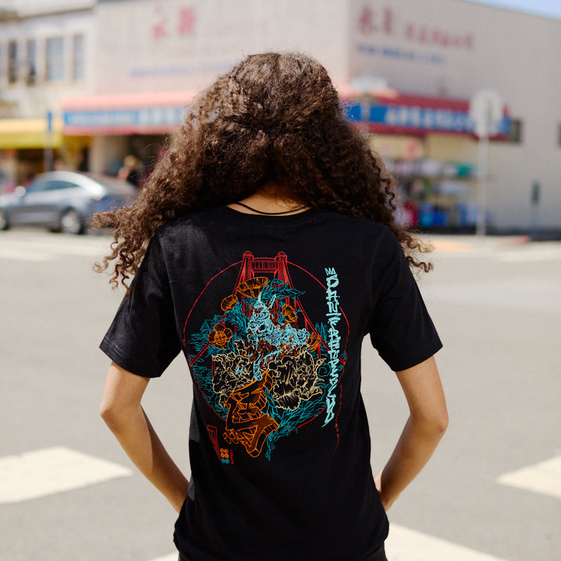 A person with curly hair wearing a Year of the Horse Tee stands at a crosswalk in a city neighborhood.