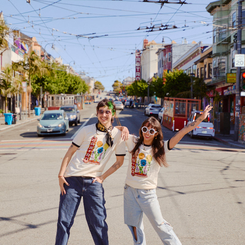 Two people in Castro Pride Tees and sunglasses pose playfully on a sunny city street near Castro Theatre, their vibrant shirts standing out amid the lively city backdrop.