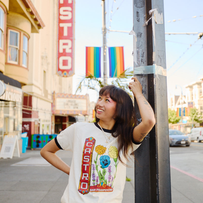 A smiling woman in a Castro Pride Tee stands on a city street with the Castro Theatre sign and a rainbow flag behind her, capturing the spirit of Pride.