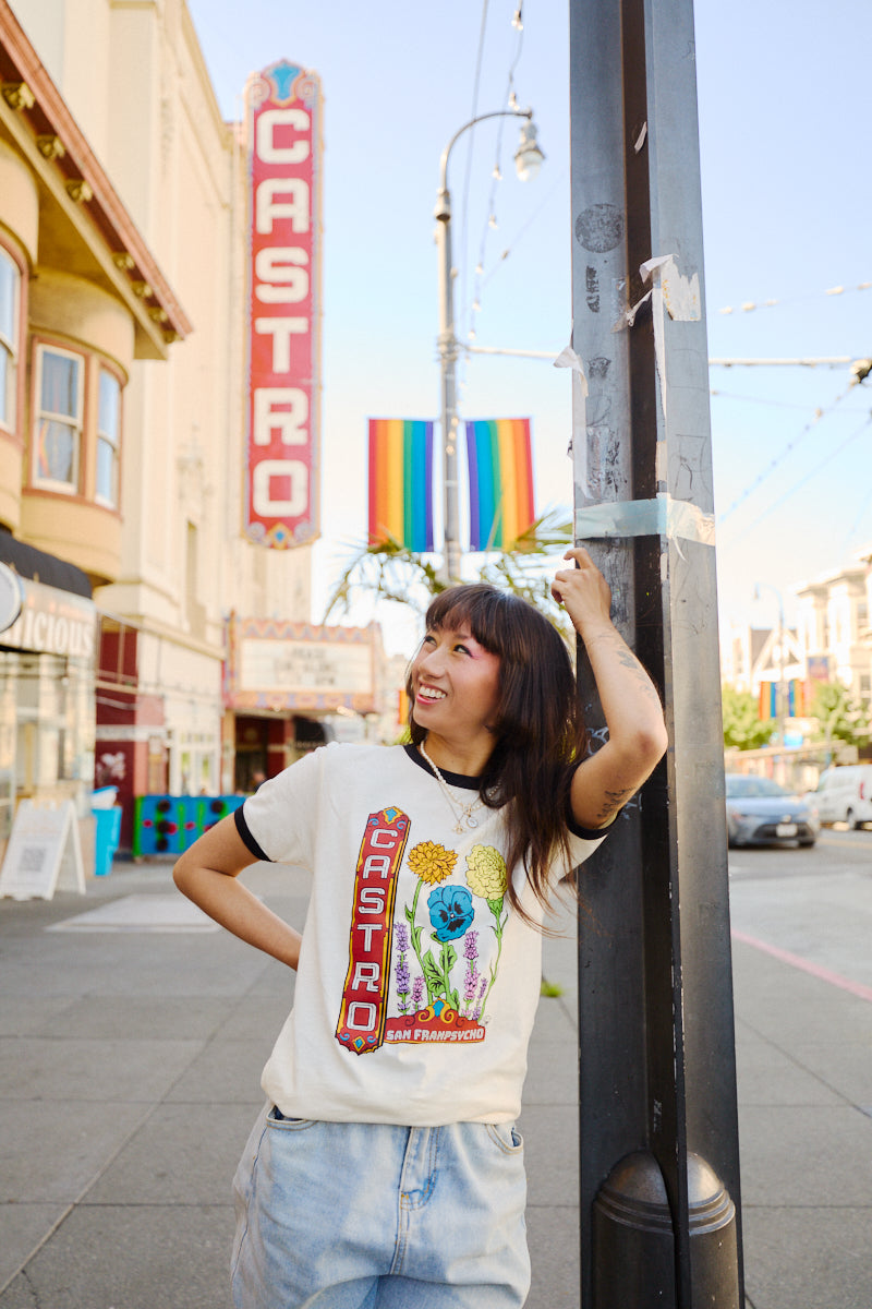 A smiling woman in a Castro Pride Tee stands on a city street with the Castro Theatre sign and a rainbow flag behind her, capturing the spirit of Pride.