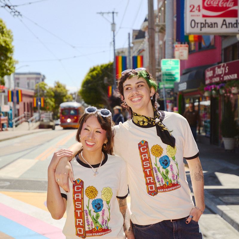 Two smiling people wearing Castro Pride Tees stand on a sunny, rainbow-striped street in San Francisco, showcasing the shirt's bold Pride design.