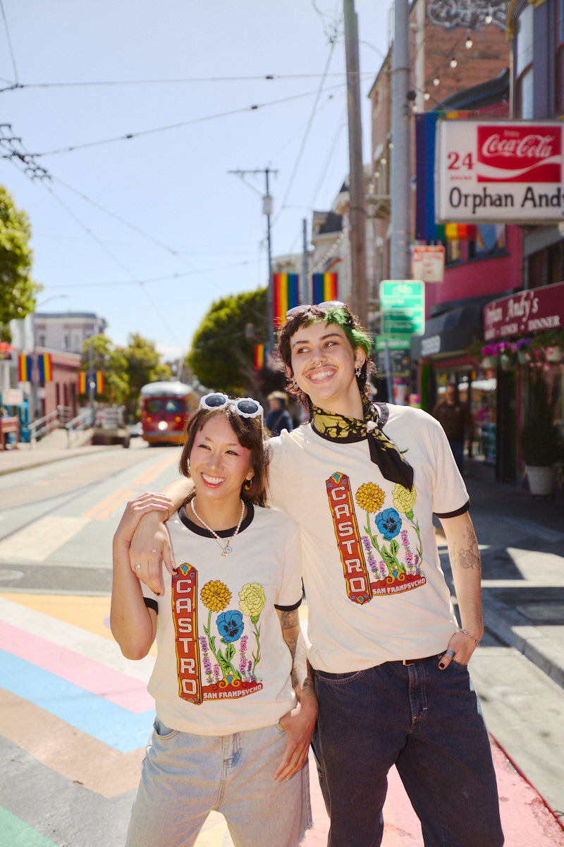 Two smiling people wearing Castro Pride Tees stand on a sunny, rainbow-striped street in San Francisco, showcasing the shirt's bold Pride design.