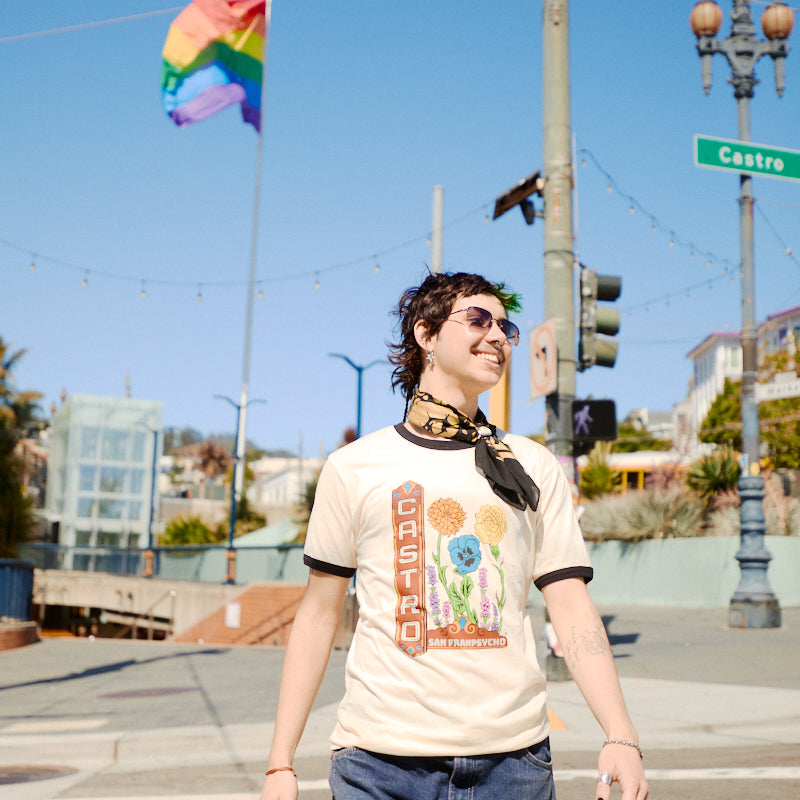 A person wearing the Castro Pride Tee smiles on a sunny street, rainbow flag and iconic Castro Theatre sign in the background, celebrating with queer flowers.