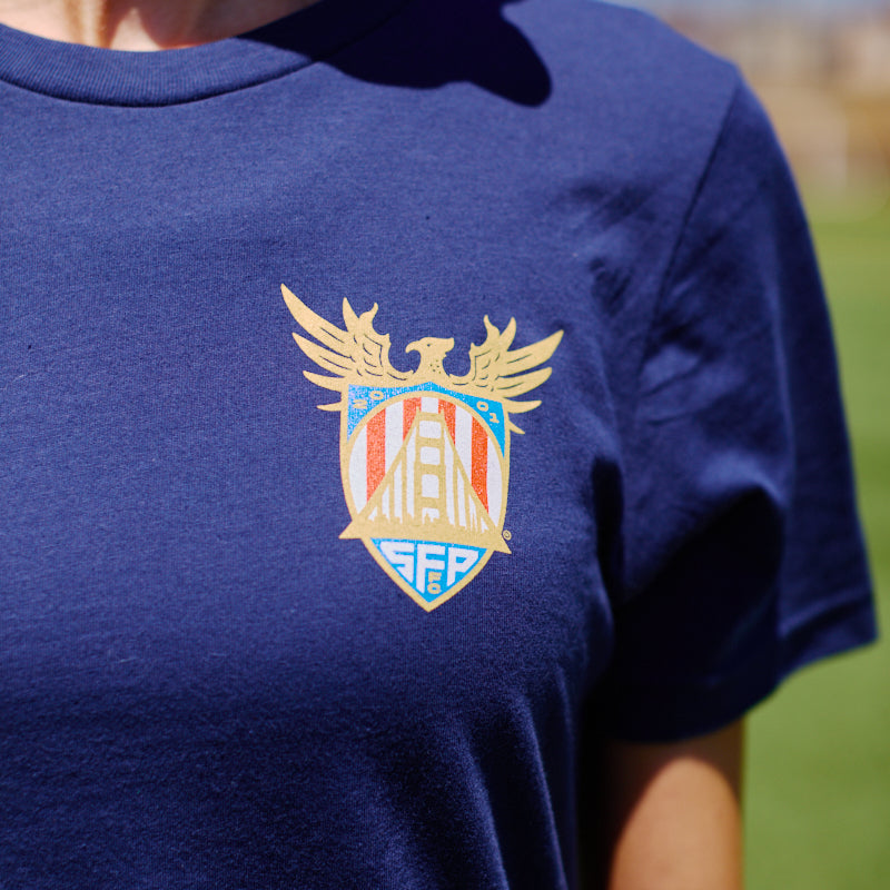 Wearing the WC x SFP Tee—a navy shirt featuring a vibrant, winged crest on the chest—soak up the sun and show off your Team USA pride outdoors.