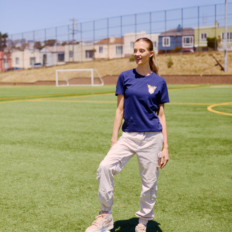 Woman on a soccer field in San Francisco, one foot on a blue and white soccer ball, goalposts behind her—sporting the WC x SFP Tee and ready to represent Team USA at the World Cup.