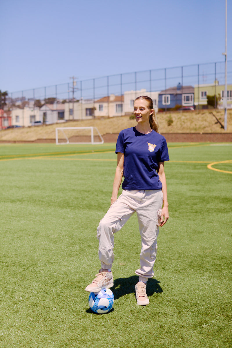 Woman on a soccer field in San Francisco, one foot on a blue and white soccer ball, goalposts behind her—sporting the WC x SFP Tee and ready to represent Team USA at the World Cup.