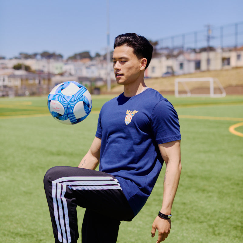 Wearing the WC x SFP Tee, a man balances a blue and white soccer ball on his knee in sunny San Francisco, channeling Team USA’s energy as he trains for the World Cup.