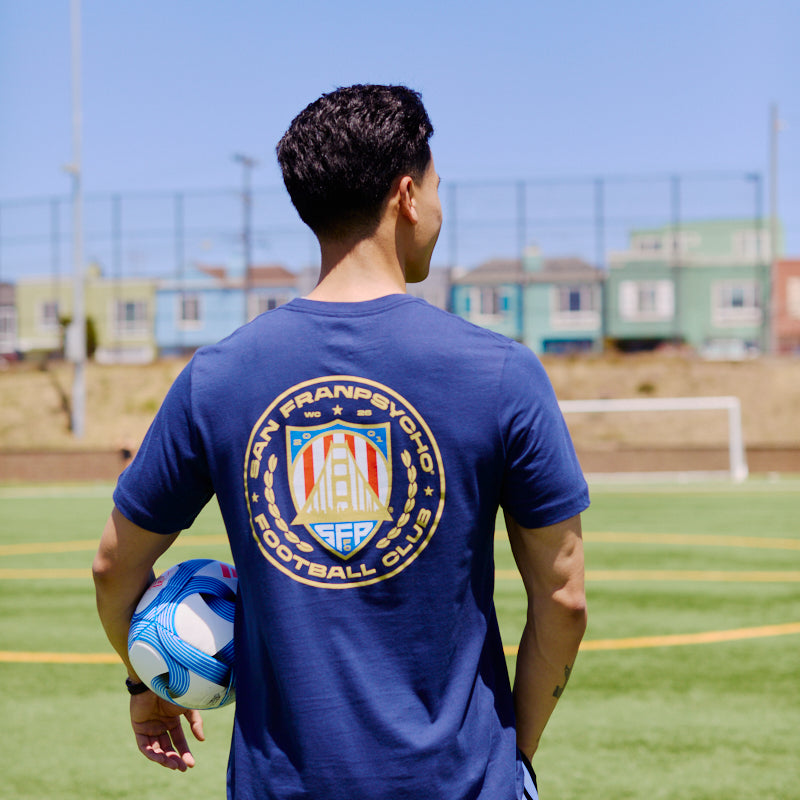 Man on a soccer field holding a ball, wearing the WC x SFP Tee in blue with a Team USA football club logo on the back.