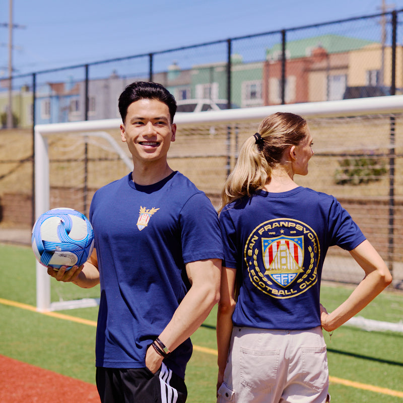 Two people are on a soccer field in San Francisco; one holds a ball and smiles, while the other faces away, proudly showing the WC x SFP Tee.