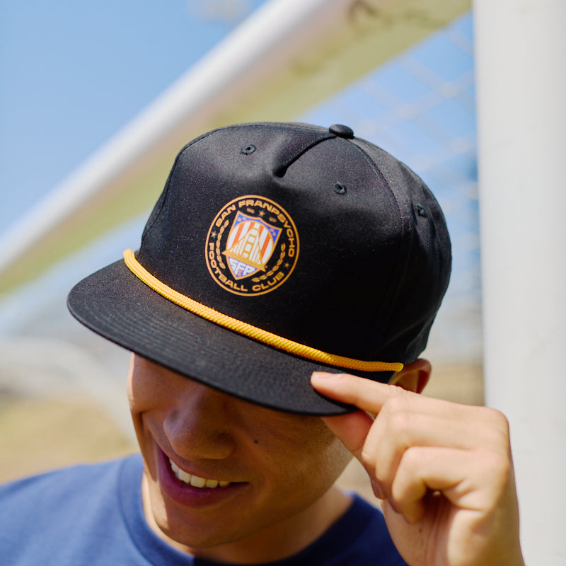 A smiling person in a blue shirt tilts the WC x SFP Hat, which features a black design with a golden emblem and trim, while standing outdoors.