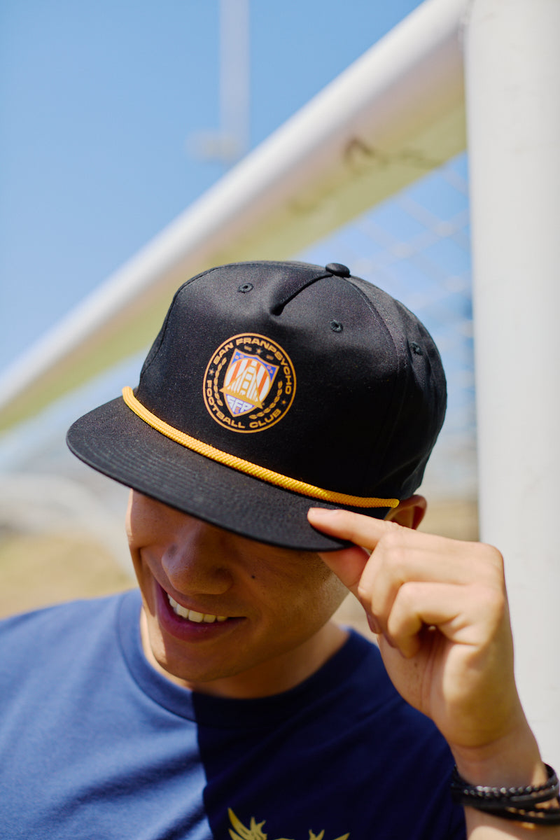 A smiling person in a blue shirt tilts the WC x SFP Hat, which features a black design with a golden emblem and trim, while standing outdoors.