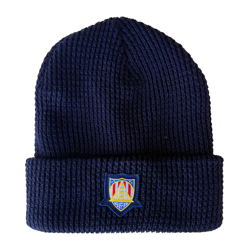 The WC x SFP Beanie is a navy blue knit hat with a folded brim and a vibrant embroidered crest, inspired by SFP beanies worn by US Men's National Team fans.