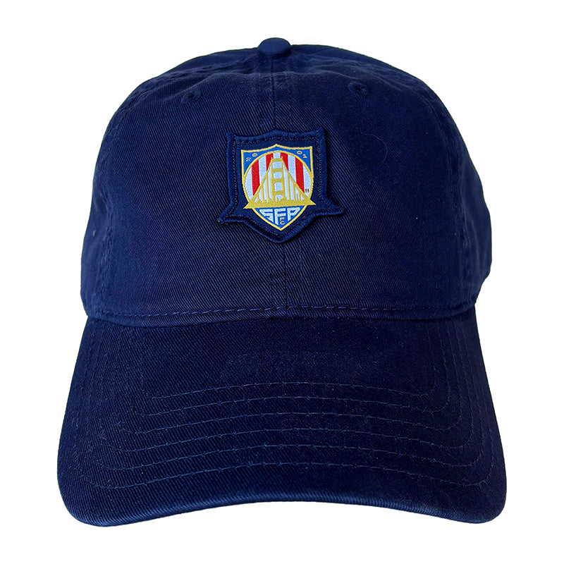 The WC x SFP Patched Hat is a navy blue baseball cap featuring a colorful embroidered shield logo on the front, perfect for any US Men's National Team or World Cup fan.