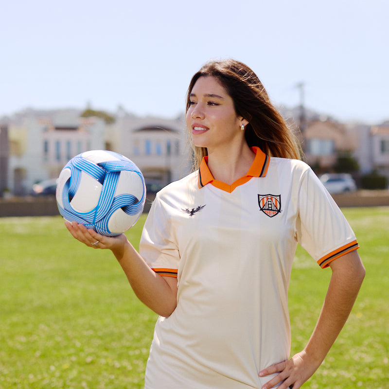A woman wearing the WC x SFP Jersey - Away holds a blue and white soccer ball on a grassy field.