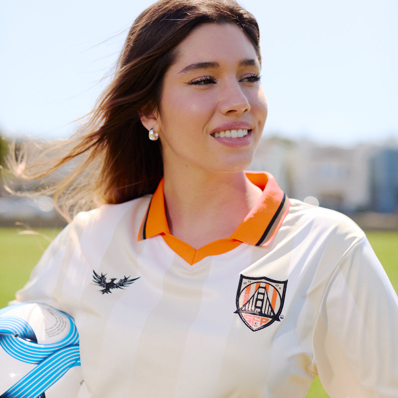 A woman wearing the WC x SFP Jersey - Away holds a blue and white soccer ball while standing on a grassy field.