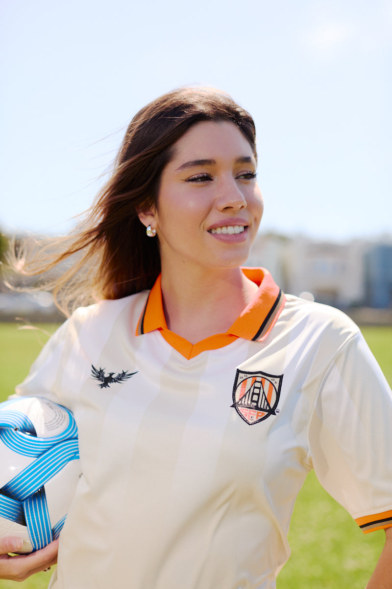 A woman wearing the WC x SFP Jersey - Away holds a blue and white soccer ball while standing on a grassy field.