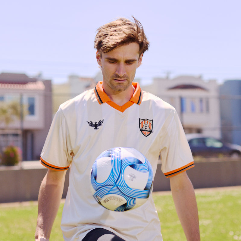A man wearing the WC x SFP Jersey - Away juggles a blue and white soccer ball outdoors on a sunny day.
