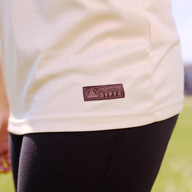 Person wearing the WC x SFP Jersey - Away, a cream shirt with a small patch and fan fit style, paired with black leggings, standing on grass outdoors.