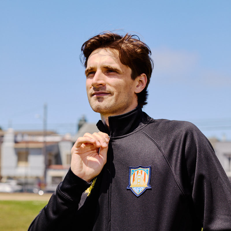 A man wears a WC x SFP Zip-Up Jacket with a sports logo while standing outdoors on a sunny day.