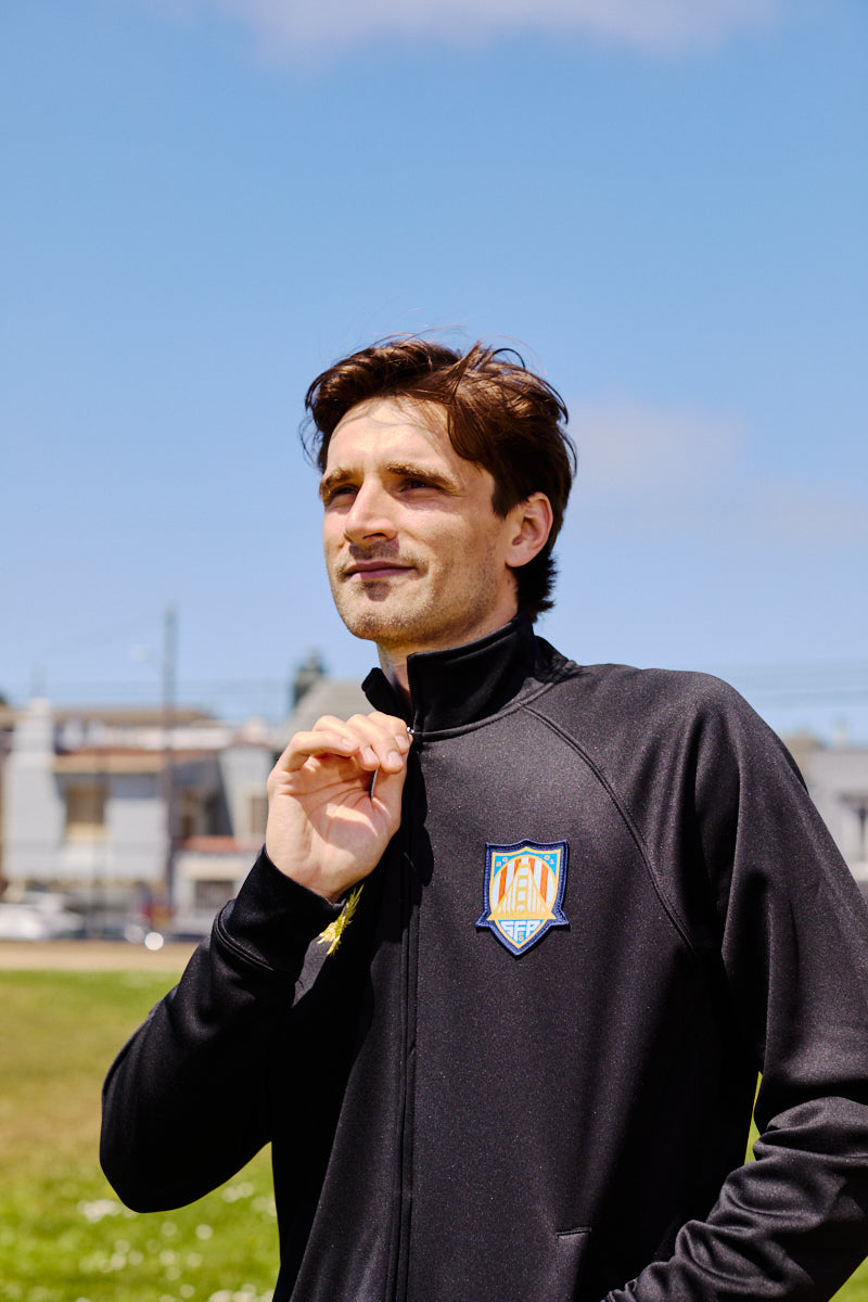 A man wears a WC x SFP Zip-Up Jacket with a sports logo while standing outdoors on a sunny day.