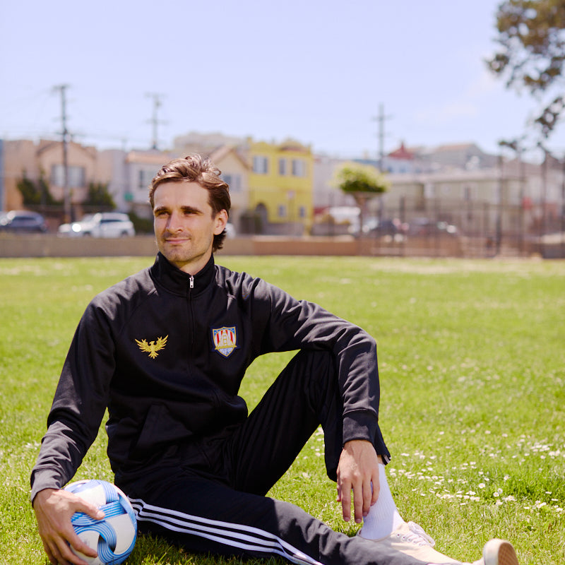 A man wearing the WC x SFP Zip-Up Jacket sits on grass with a soccer ball under the sun, capturing the US Men's National Team spirit in a park.