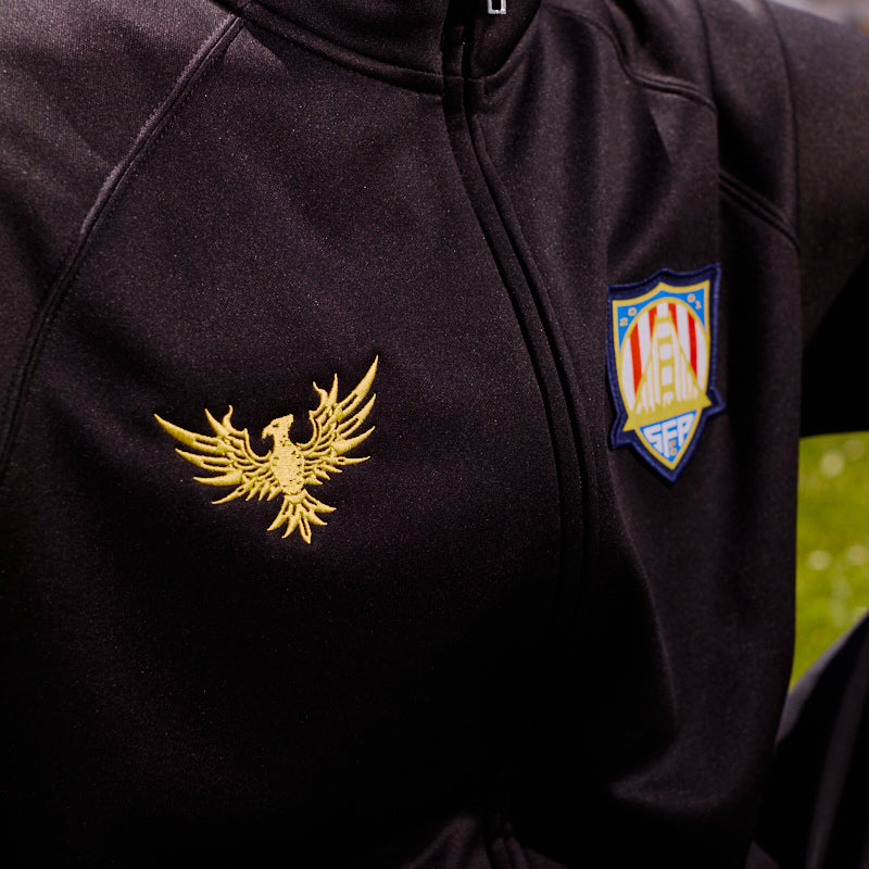Wearing the WC x SFP Zip-Up Jacket—a black zip-up with a gold eagle emblem and a striking San Francisco Rising Phoenix shield patch on the chest.