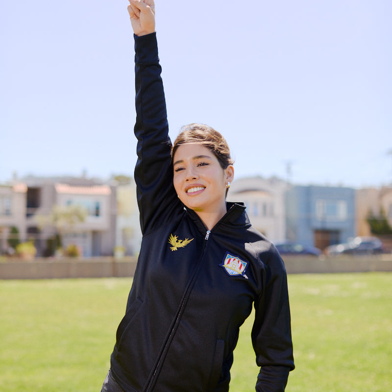 Woman standing on grass, smiling and raising one arm with index finger pointing to the sky, wearing the WC x SFP Zip-Up Jacket.