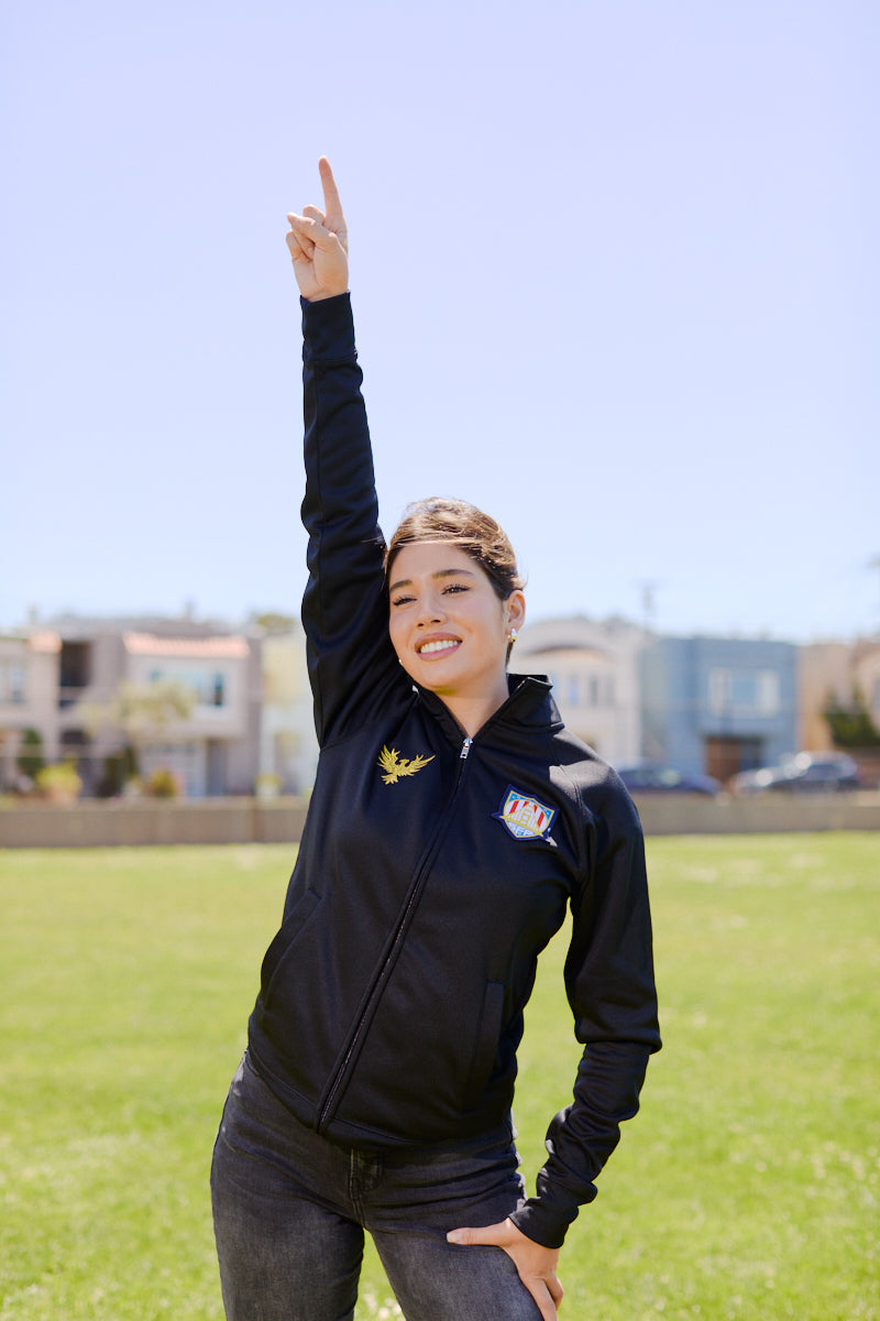 Woman standing on grass, smiling and raising one arm with index finger pointing to the sky, wearing the WC x SFP Zip-Up Jacket.