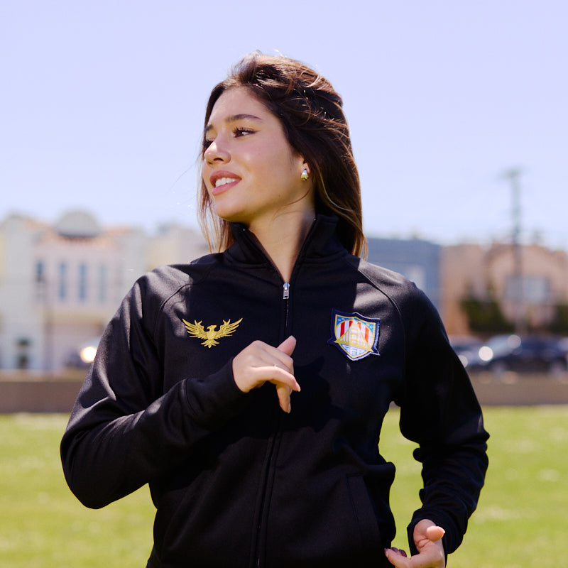A woman runs outdoors on a sunny day wearing the WC x SFP Zip-Up Jacket, with city buildings and green grass in the background.