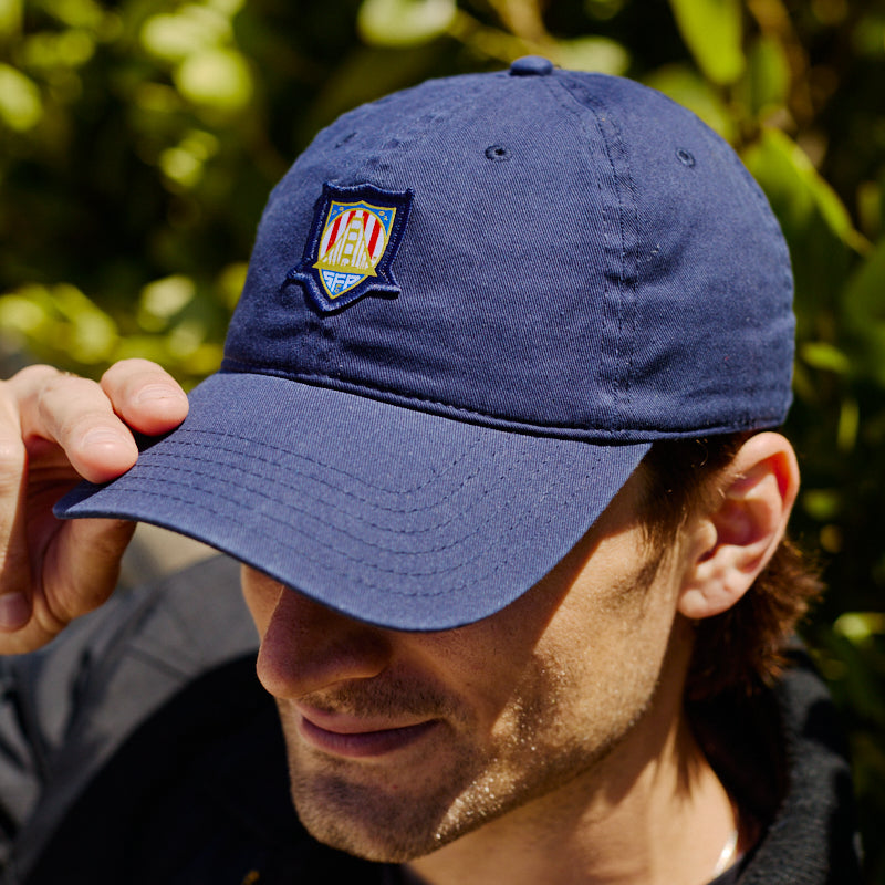 A man outdoors adjusts the brim of a blue WC x SFP Patched Hat, which features a colorful embroidered patch.