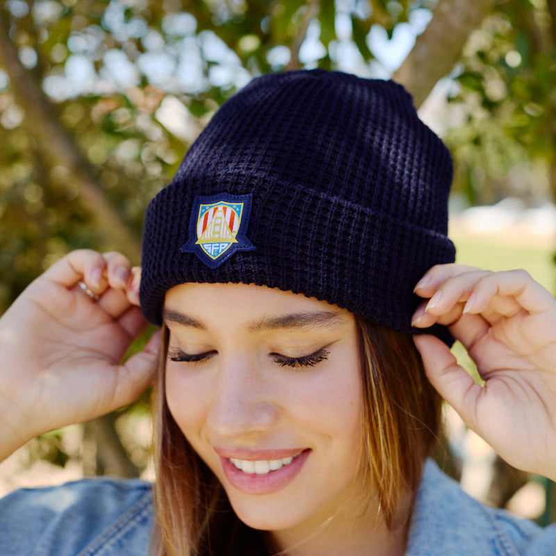 A woman smiles outdoors, wearing the WC x SFP Beanie with a colorful patch and a light denim jacket—the perfect look for cheering on the US Men's National Team.