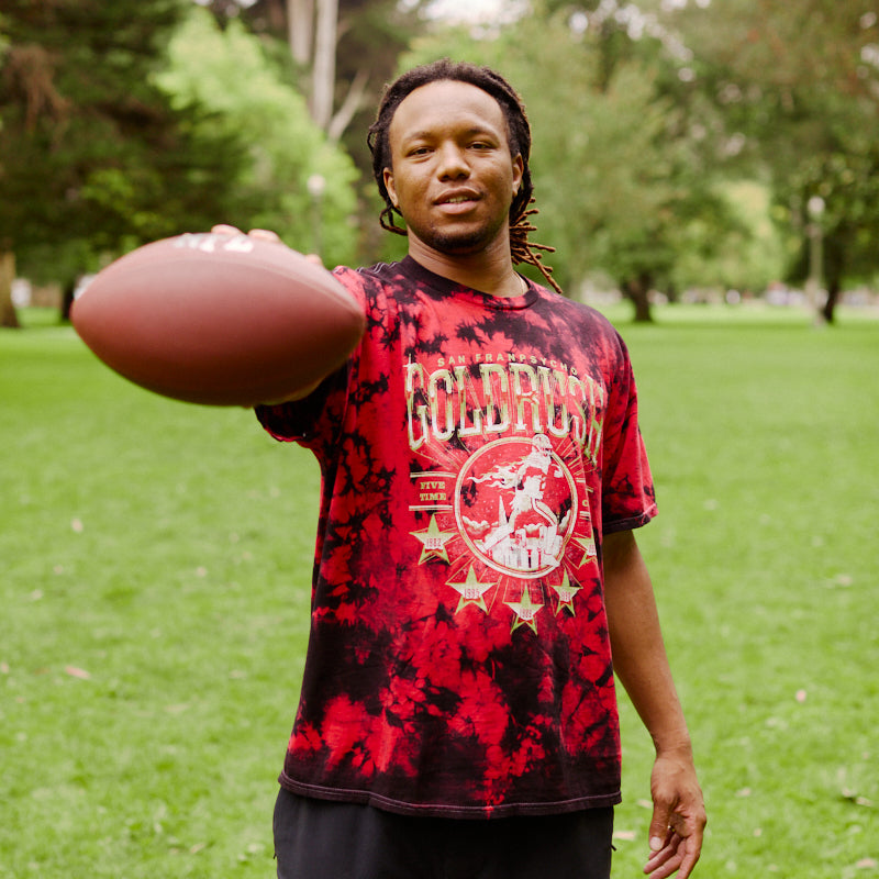 A man stands on grass in a park, holding a football out in front of him, wearing the Gold Rush Tee - Red Crystal Dye.