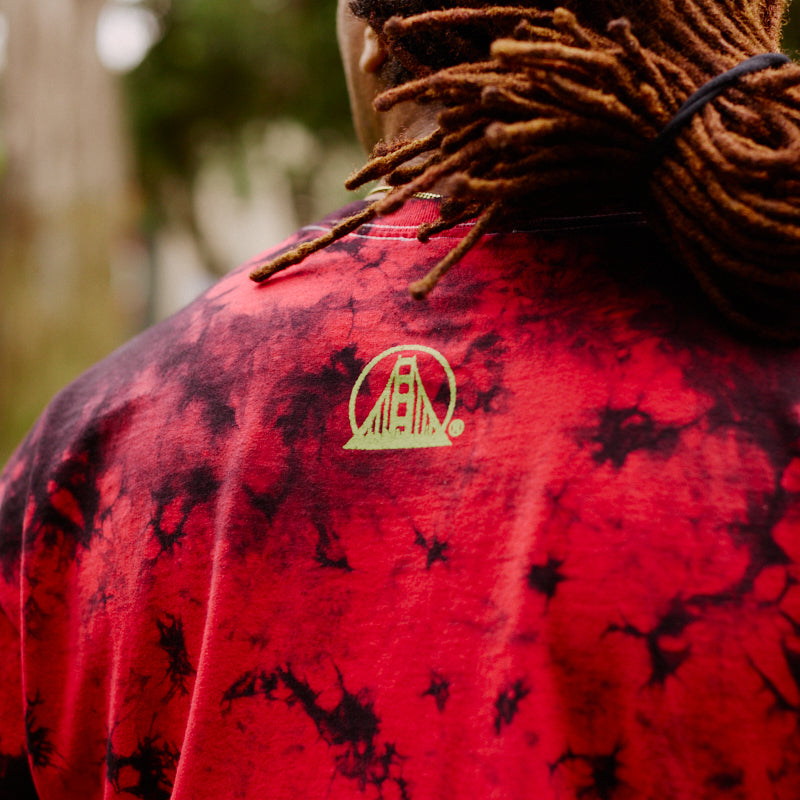 A person with dreadlocks wears the Gold Rush Tee - Red Crystal Dye, a unisex red tie-dye shirt featuring a yellow bridge logo on the back, outdoors—perfect for any 49ers or Gold Rush fan.