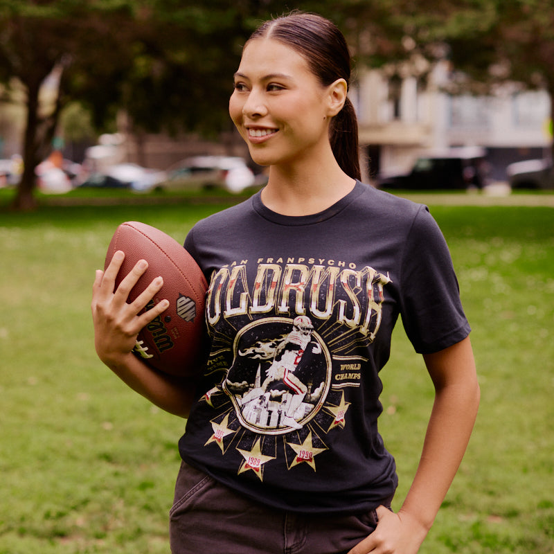 Woman smiling while holding a football in one hand, standing on a grassy field in a park, wearing the Gold Rush Tee in Vintage Black.