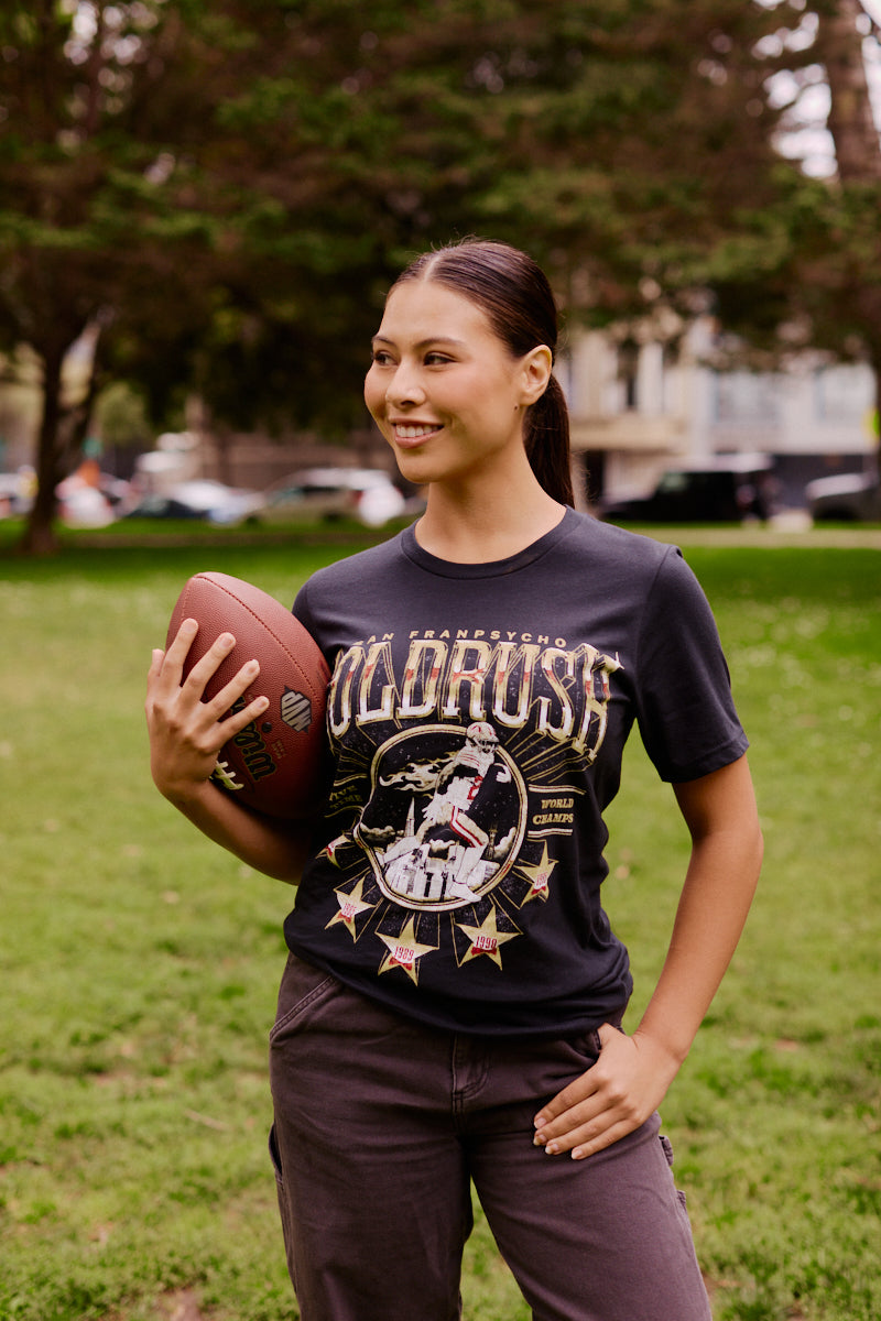 Woman smiling while holding a football in one hand, standing on a grassy field in a park, wearing the Gold Rush Tee in Vintage Black.