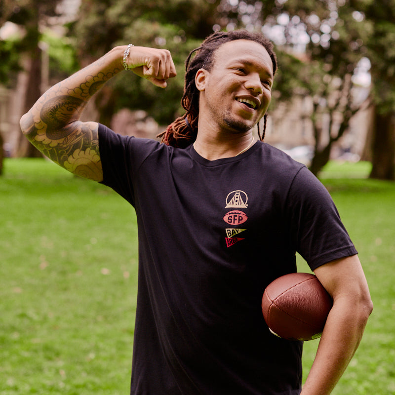 With a football tucked under one arm and his other arm flexed in the park, a smiling man with dreadlocks proudly sports the Niners Faithful Tee—perfect for showing off that San Francisco spirit.