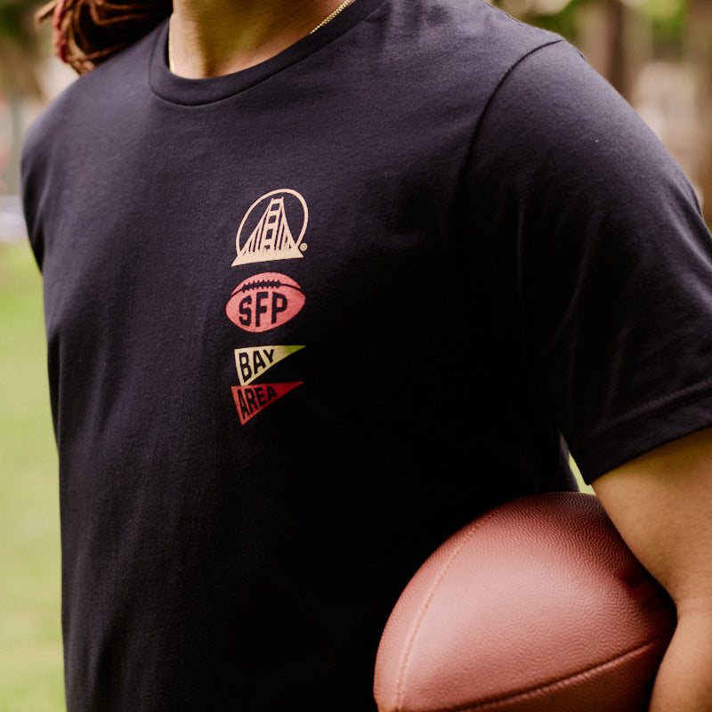 A person stands outdoors on the grass, holding a football under their arm and wearing the Niners Faithful Tee—a unisex black shirt perfect for showing off your San Francisco pride.
