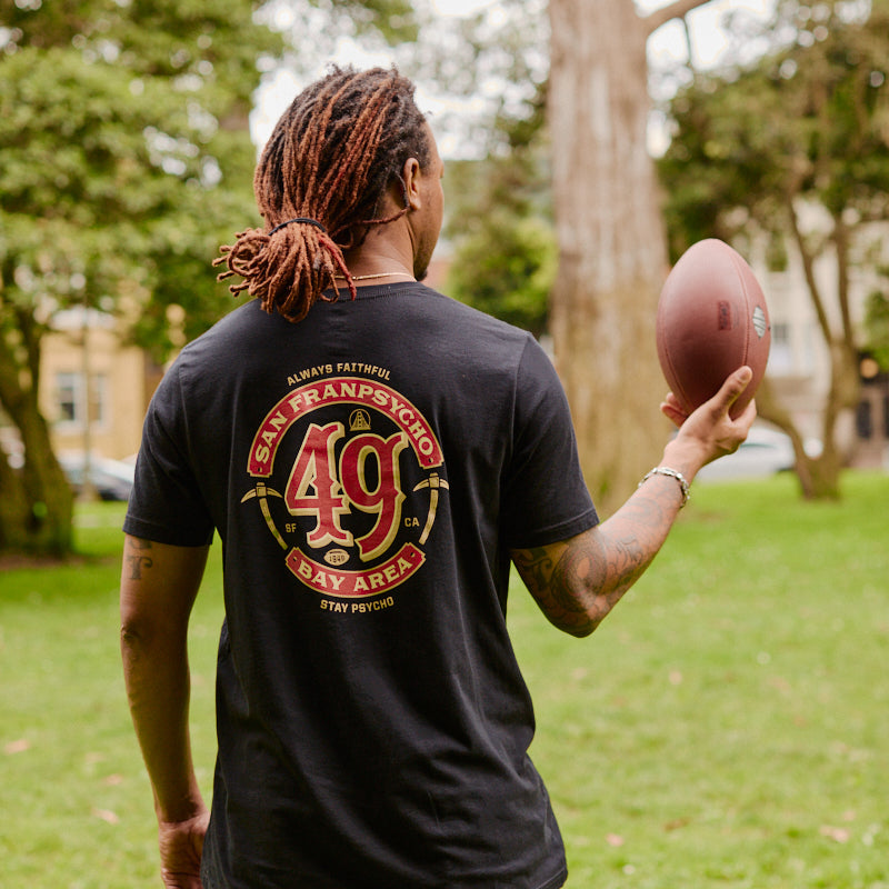 Wearing the Niners Faithful Tee, a person with dreadlocks holds a football while standing in a grassy park in San Francisco.