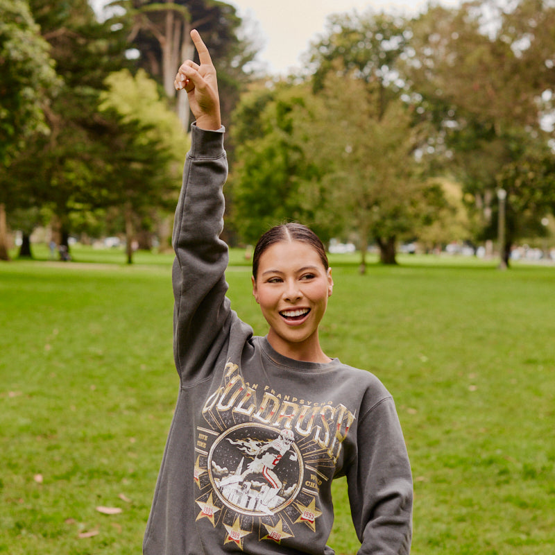 A smiling person wearing the Gold Rush Crewneck stands on grass in a park, raising one arm and pointing upward.