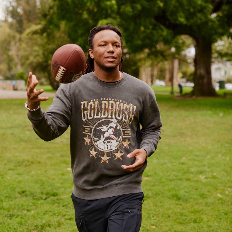 A man wearing the Gold Rush Crewneck tosses a football while walking on green grass in a park.