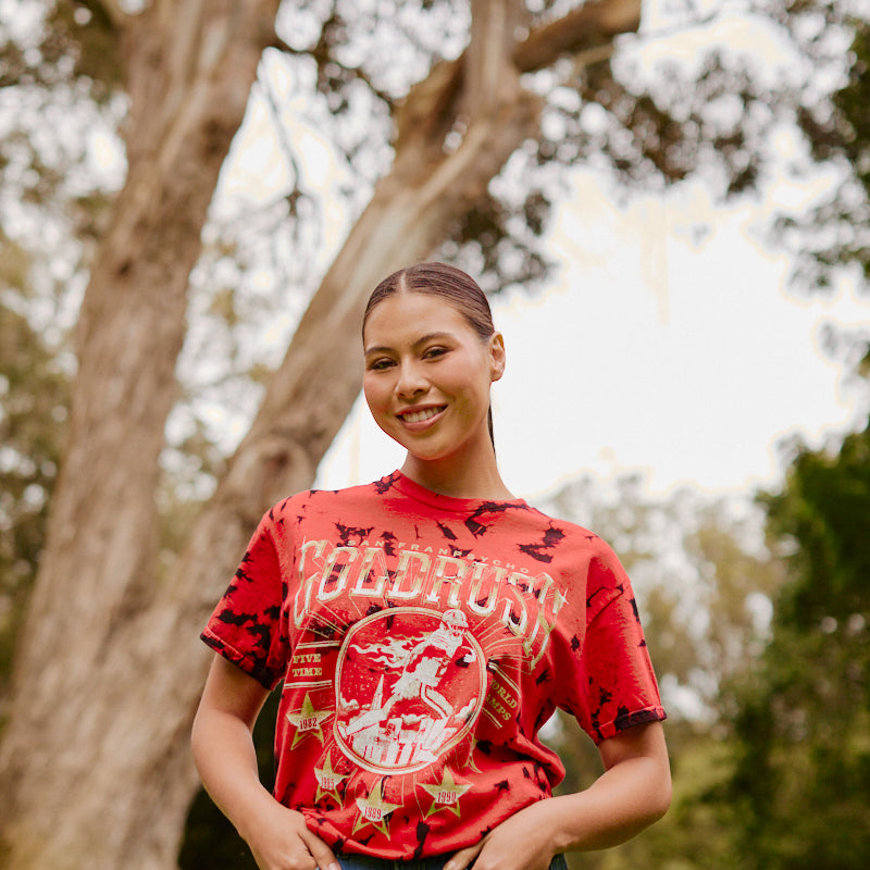 A woman smiling outdoors in the Gold Rush Tee - Red Crystal Dye and jeans, standing in front of trees in a park.