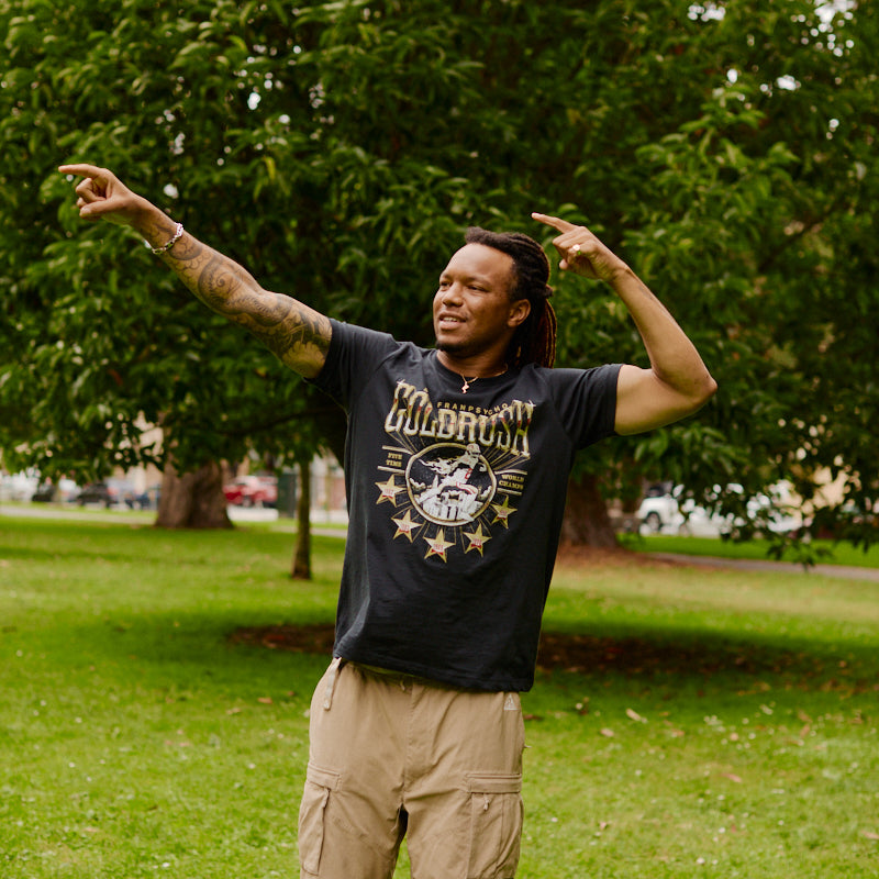 A person stands on grass, pointing to the side and looking up, wearing the Gold Rush Tee in Vintage Black with trees and greenery in the background—a classic San Francisco style.