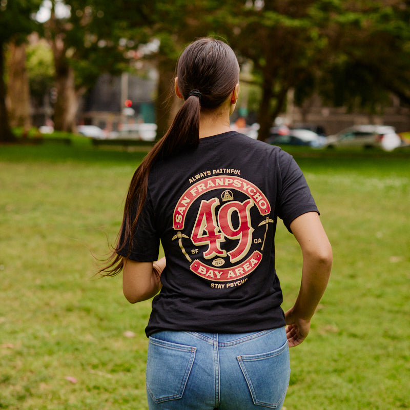 A woman with a ponytail walks on grass, wearing the Niners Faithful Tee with a classic unisex fit; trees are visible in the background.