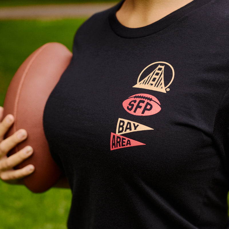 Person holding a football, wearing the Niners Faithful Tee—a black unisex fit shirt featuring SFP Bay Area and a bridge graphic on the front—making it an ideal addition to any San Francisco Niners fan collection.
